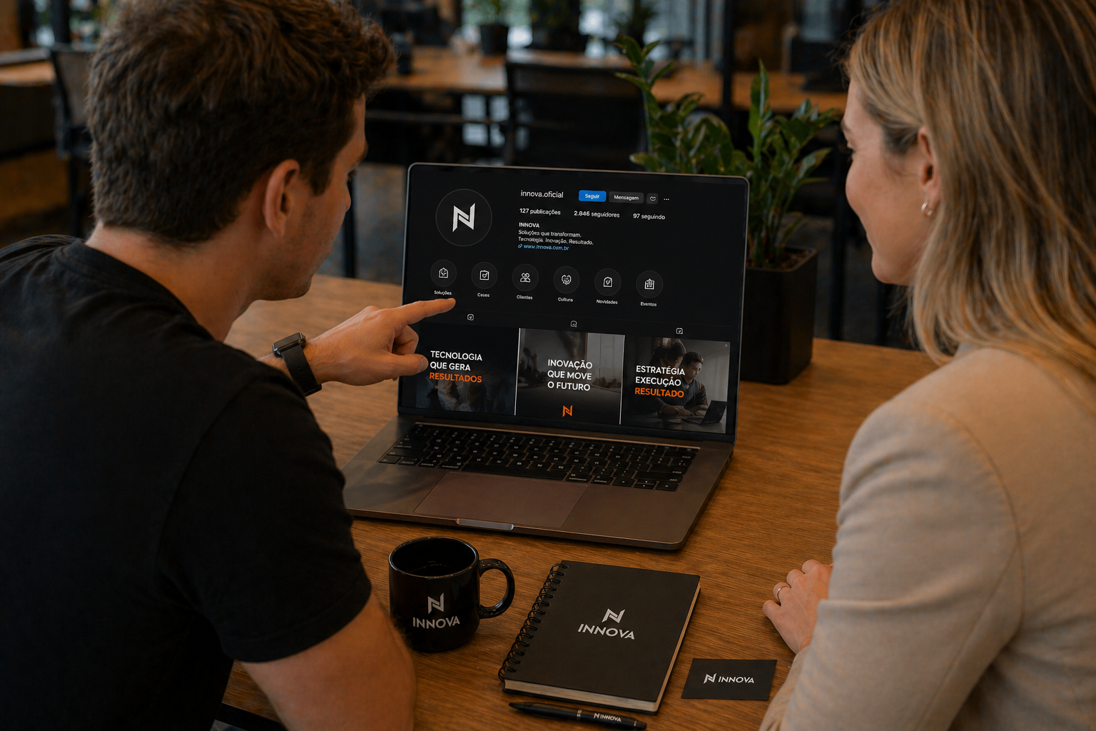 Dupla analisando o perfil da Innova nas redes sociais em um notebook, com caneca e cadernos da marca sobre a mesa